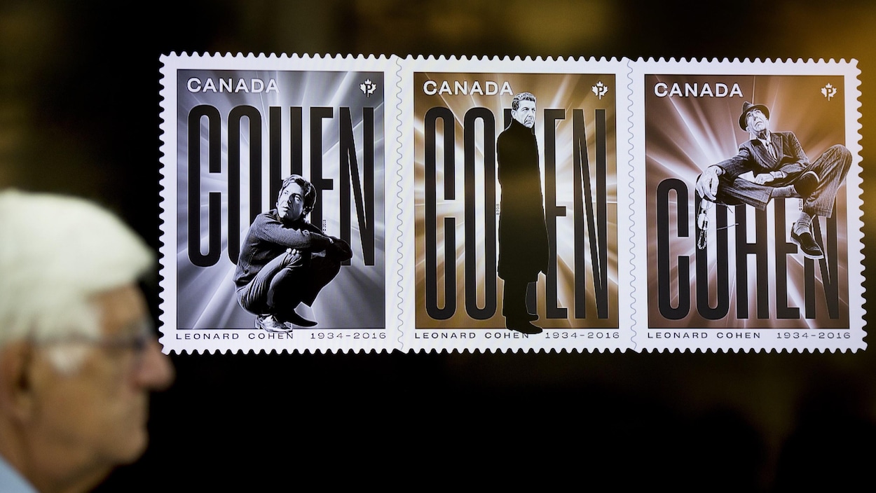 Un homme se tient à côté d'un écran où sont affichés les timbres à l'effigie de Leonard Cohen.