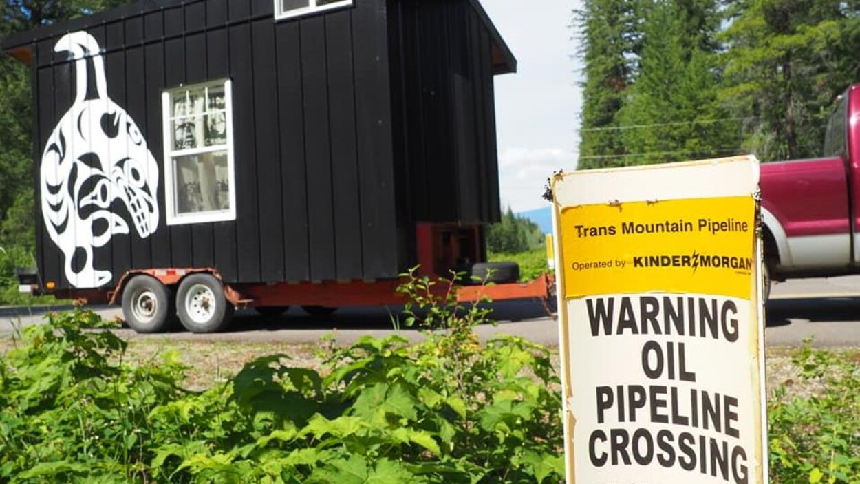 Une minuscule maison noire tirée par une camionnette avec en avant-plan une affiche où il est écrit en anglais : « Attention passage de pipeline ».