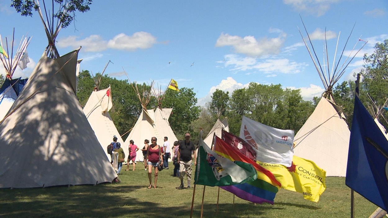 Sept tipis installés sur un gazon avec des drapeaux qui flottent autour et une dizaine de personnes qui s'y trouvent également.