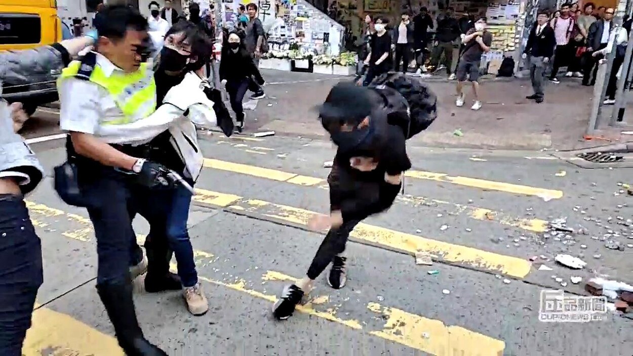 Vêtu de noir et masqué, le manifestant se tient l'abdomen, alors que deux autres protestataires s'attaquent à l'agent de police.