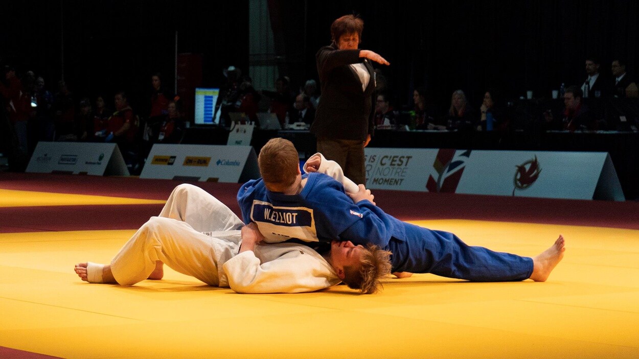 Un homme se tient couché sur un autre dans une prise de judo, avec l'arbitre qui signale le point.