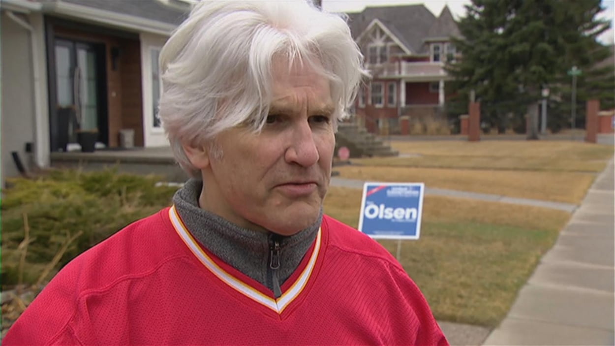 Tom Olsen, qui a des cheveux blancs et porte un chandail de hockey, parle à la caméra, dehors. Derrière lui, il y a une affiche électorale avec son nom.