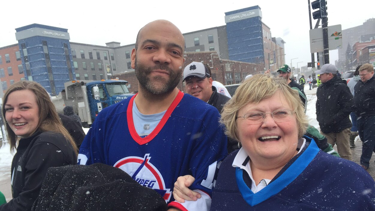 Un homme et une femme souriante portant tous deux un chandail des Jets de Winnipeg sont à l'extérieur entouré d'autres passants. Un homme et une femme souriante portant tous deux un chandail des Jets de Winnipeg sont à l'extérieur entouré d'autres passants.