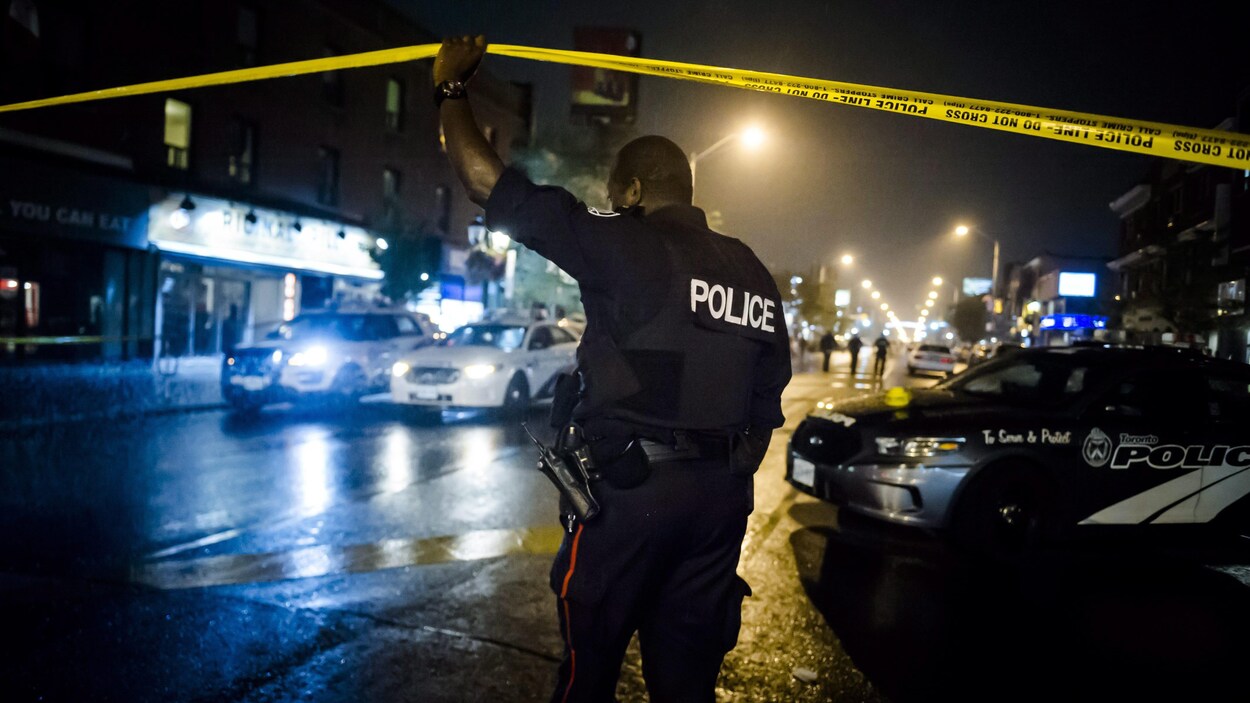 Un policier en uniforme soulève un ruban qui sert à fermer un secteur lorsque les policiers érigent un périmètre de sécurité. La photographie a été prise le soir, car il fait sombre et seuls les néons et lampadaires illuminent la scène.