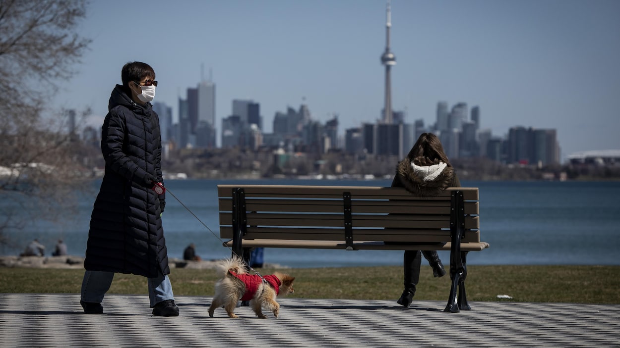 Une femme avec un masque passe à distance d'un banc public où est assis une autre femme qui regarde la ville de Toronto.
