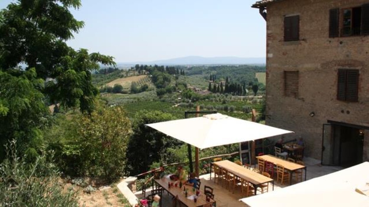 Paysage vallonneux de la Toscane, avec au premier plan une partie d'un vieux bâtiment de pierre et une terrasse sur laquelle on trouve deux grandes tables et des chaises protégées par un grand auvent.