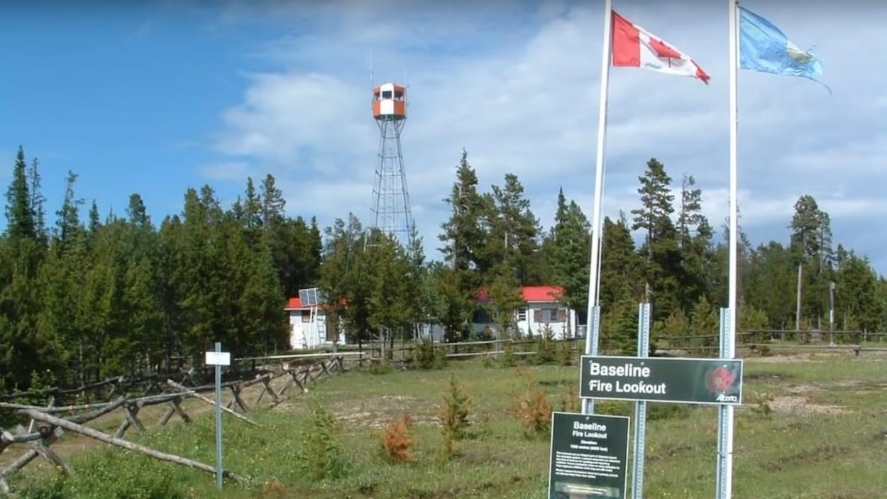 Le site d'une station de surveillance des feux. Les drapeaux canadien et albertain flottent au premier plan, tandis qu'on aperçoit la tour dépasser des arbres qui se trouvent à l'arrière-plan.