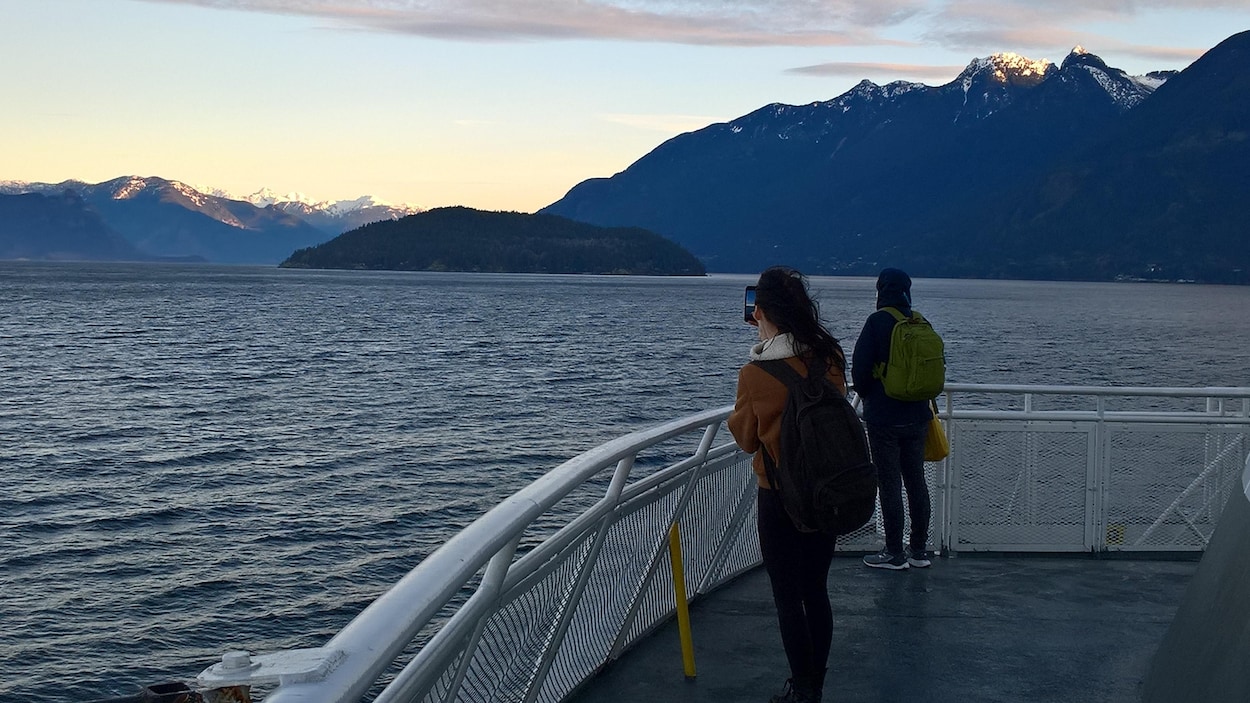 Deux passagers observent debout des montagnes enneigées depuis le pont d'un traversier qui quitte Horseshoe Bay en Colombie-Britannique.