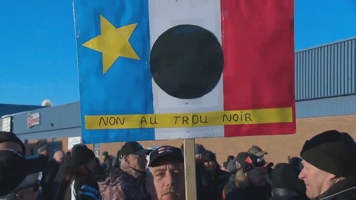 Un manifestant brandit une pancarte sur laquelle est illustré le drapeau acadien et un trou noir Un manifestant brandit une pancarte sur laquelle est illustré le drapeau acadien et un trou noir