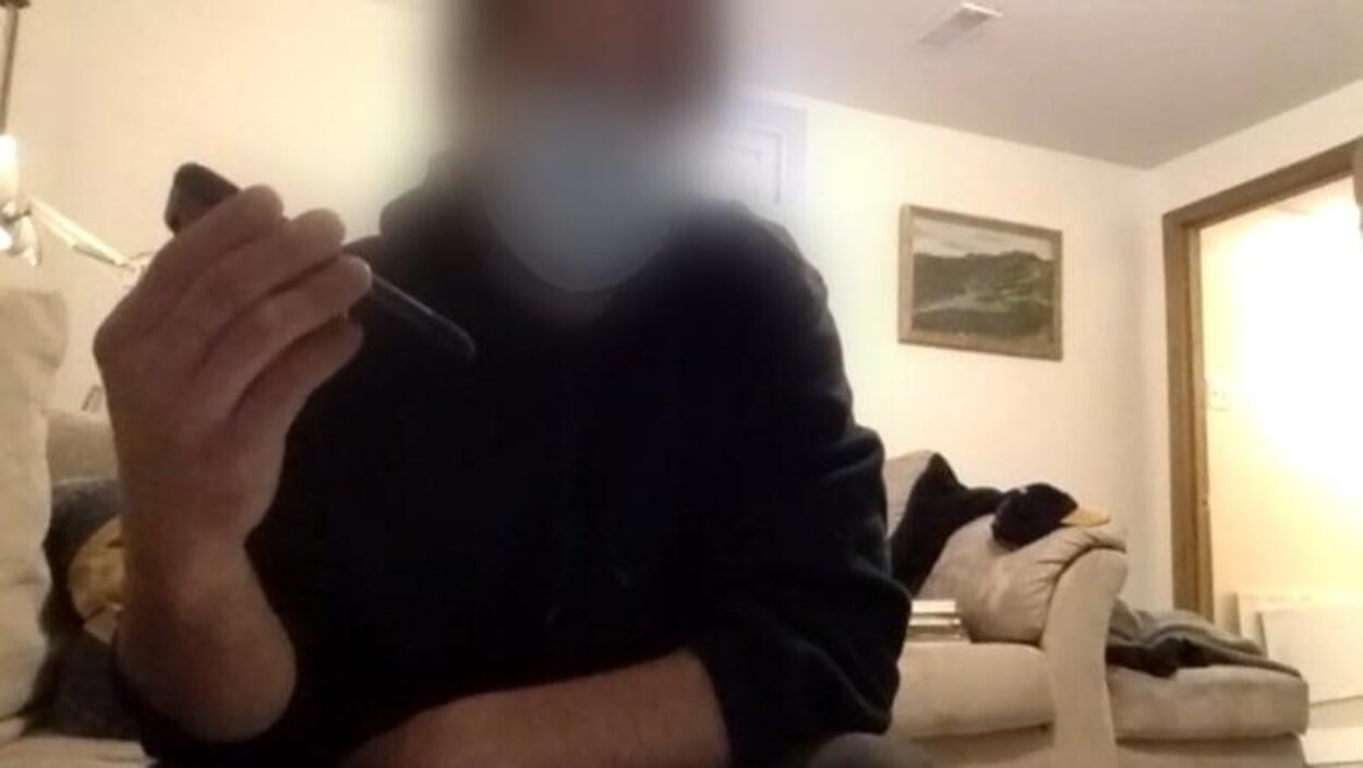 Un homme est assis dans son salon devant un écran d'ordinateur. Il a le visage brouillé.