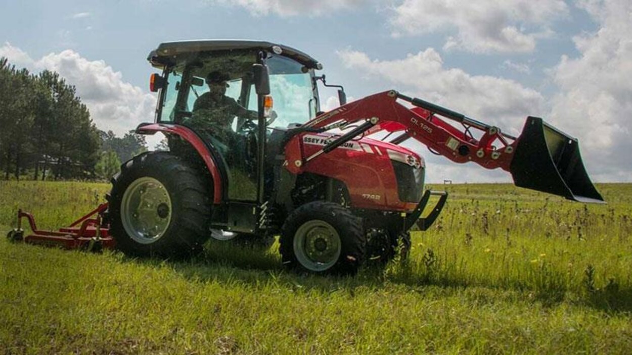 Un agriculteur dans un tracteur qui coupe l'herbe l'été dans un champ avec en arrière-plan une bordure d'arbres.