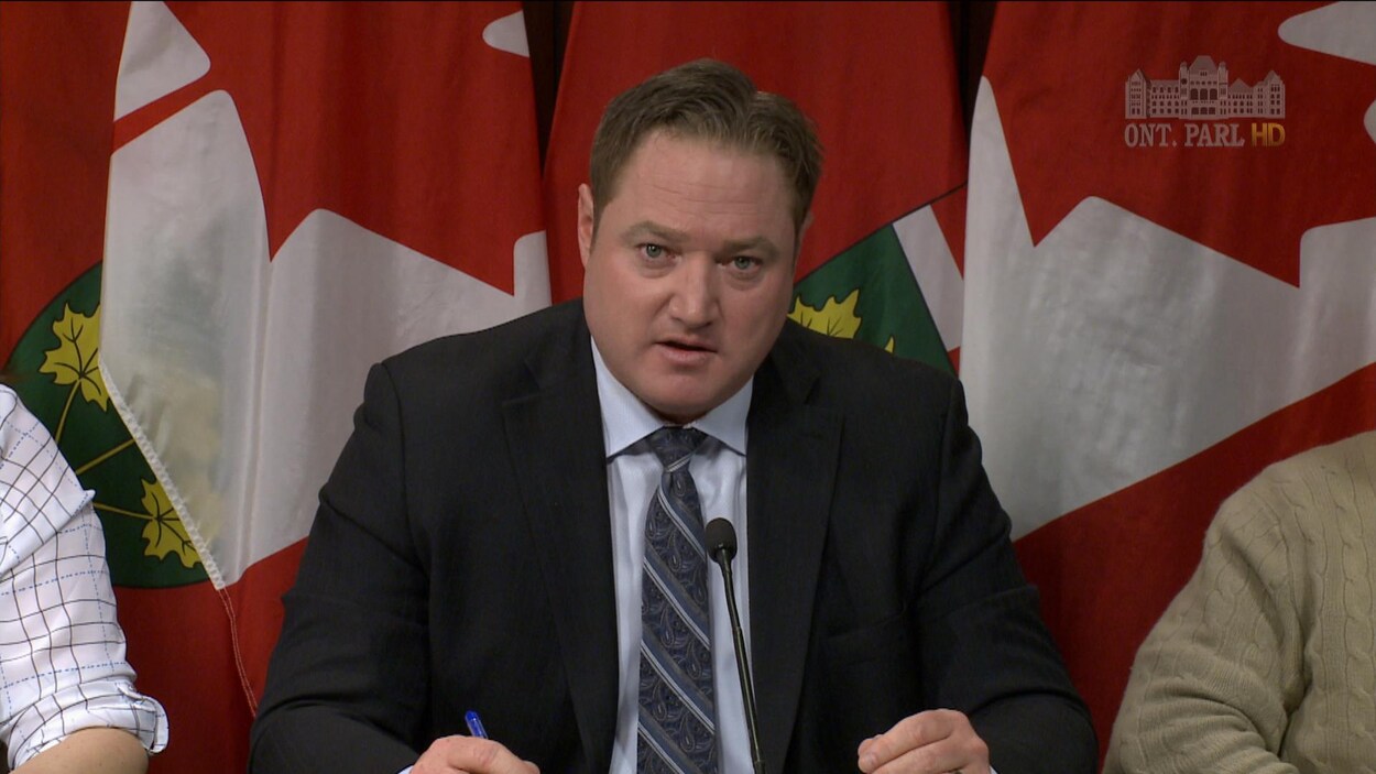 Un homme en veston-cravate parle à un micro avec des drapeaux de l'Ontario derrière lui.