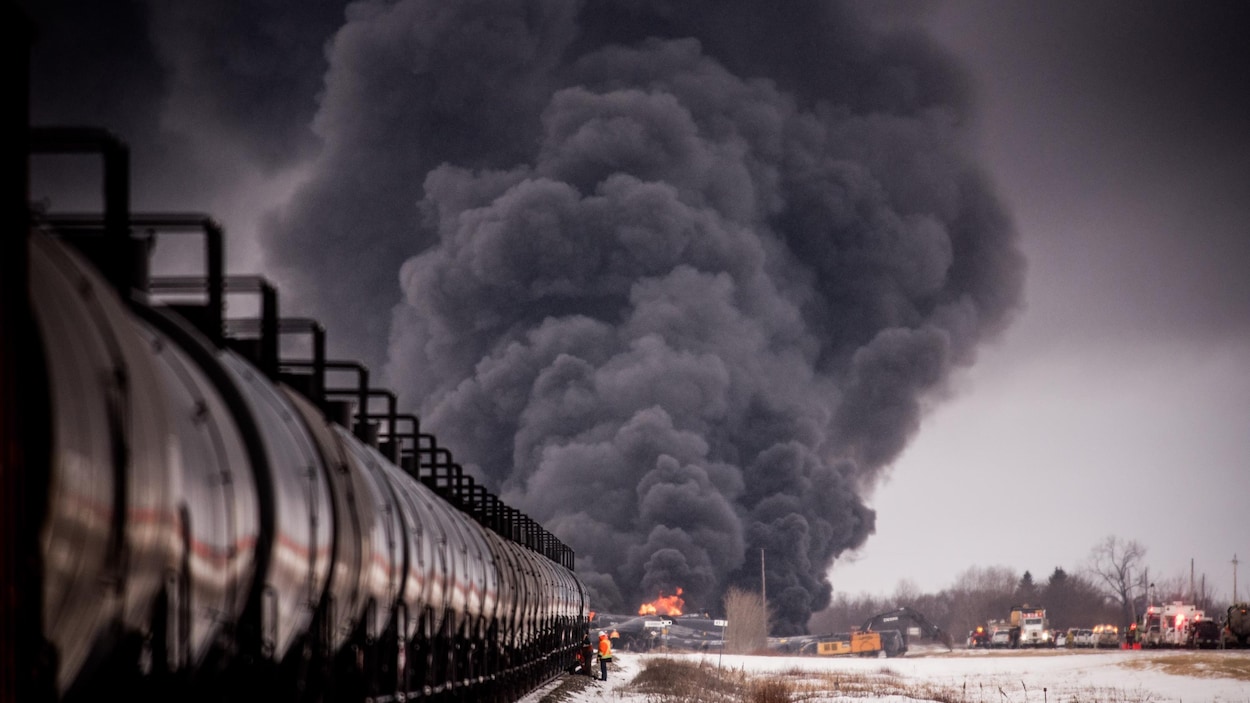 Un épais nuage de fumée se dégage d'un feu devant un long train transportant du pétrole, immobilisé sur les rails.