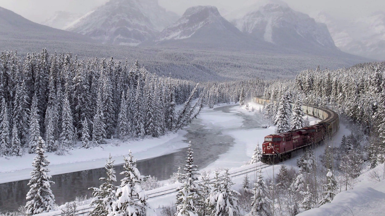 Un train rouge du Canadien Pacifique roule au milieu de sapins recouverts de neige et le long d'une rivière à moitié gelée. Au loin, on voit les sommets enneigés des montagnes Rocheuses. Un train rouge du Canadien Pacifique roule au milieu de sapins recouverts de neige et le long d'une rivière à moitié gelée. Au loin, on voit les sommets enneigés des montagnes Rocheuses.