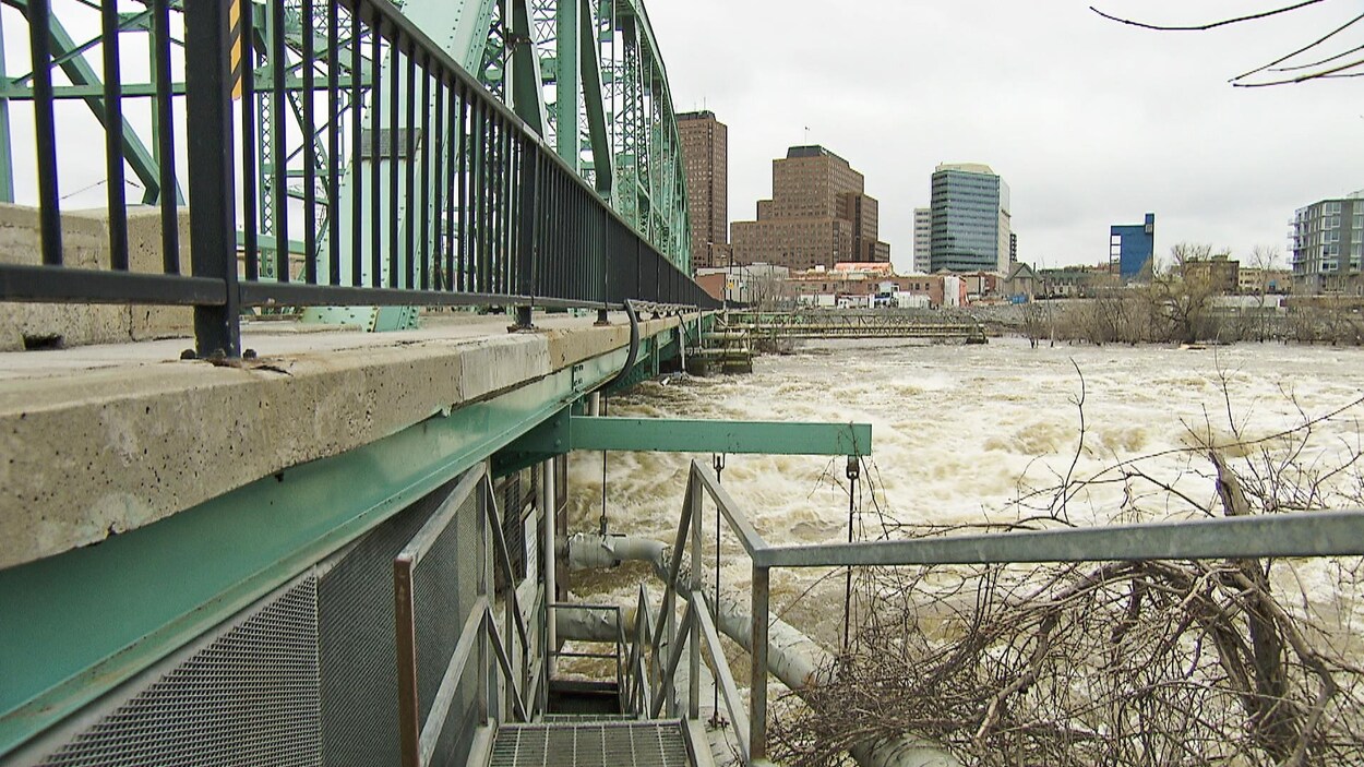 Un pont vert métallique sous lequel la rivière gronde. Vue sur le centre-ville de Gatineau.