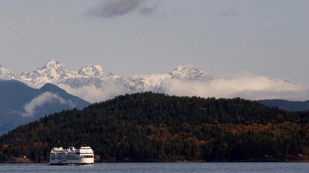 Un traversier de BC Ferries vogue en mer près de Vancouver, des montagnes de conifères et des montagnes enneigées à l'arrière-scène.