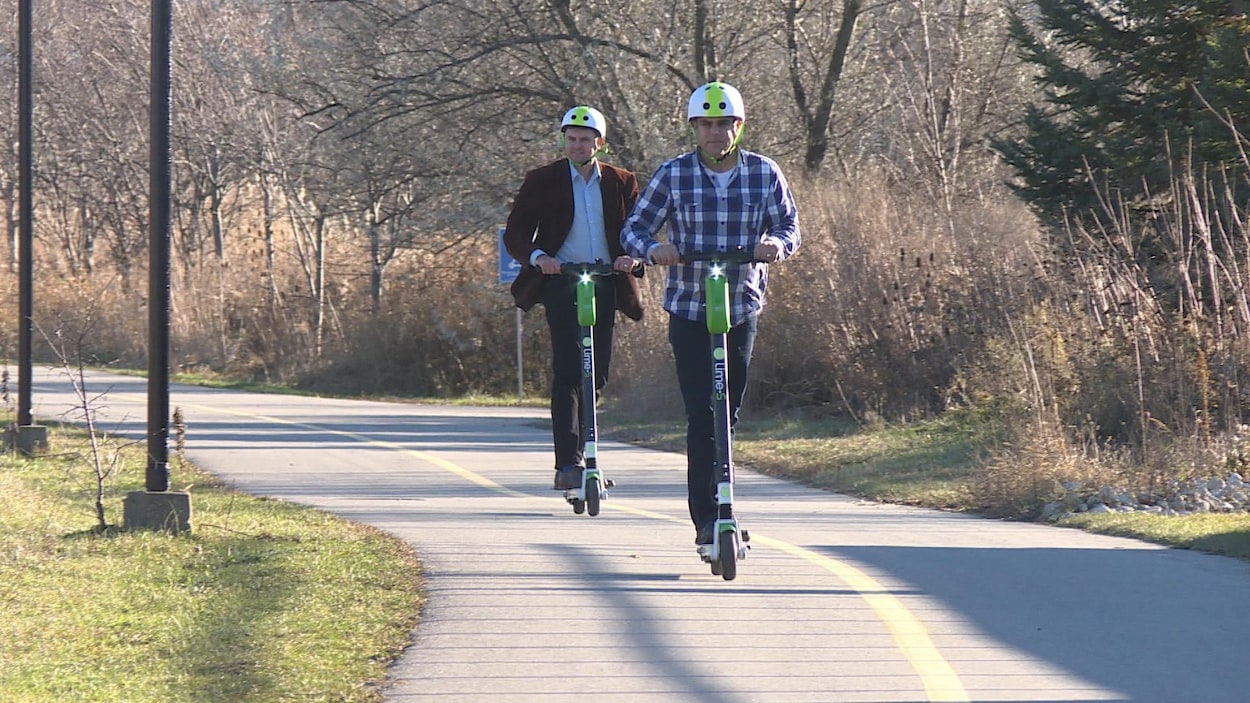 Deux hommes portant des casques, circulant sur une piste cyclable sur leur trottinettes. Ils sont l'un à l'arrière de l'autre. On peut voir de la végétation des deux côtés de la route pavée.