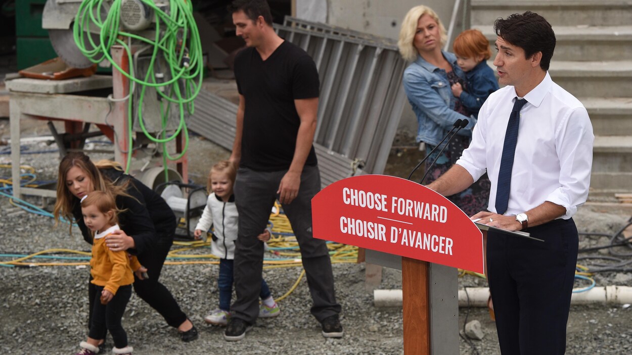 Deux femmes et un homme sont près de jeunes enfants derrière Justin Trudeau, qui parle devant un lutrin.
