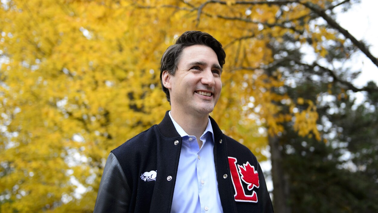 Justin Trudeau souriant, devant un arbre aux feuilles orangées.