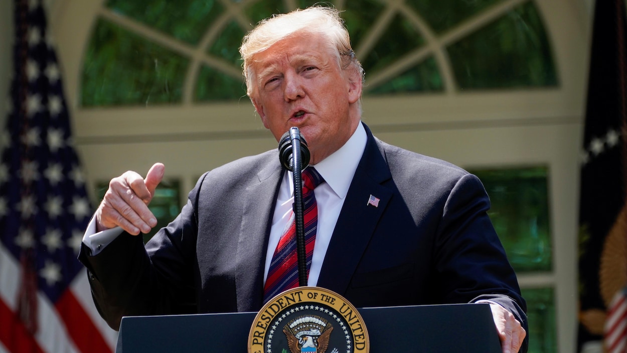Donald Trump, la main droite levée, devant un podium, où on peut lire « président des États-Unis, dans la roseraie de la Maison-Blanche ».