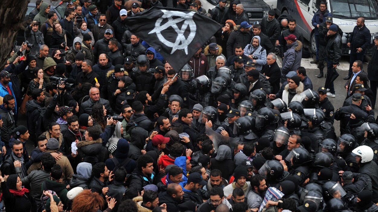 Des dizaines de manifestants affrontent des policiers, l'un d'eux tenant un drapeau noir sur lequel est peint le symbole de l'anarchie. Des dizaines de manifestants affrontent des policiers, l'un d'eux tenant un drapeau noir sur lequel est peint le symbole de l'anarchie.