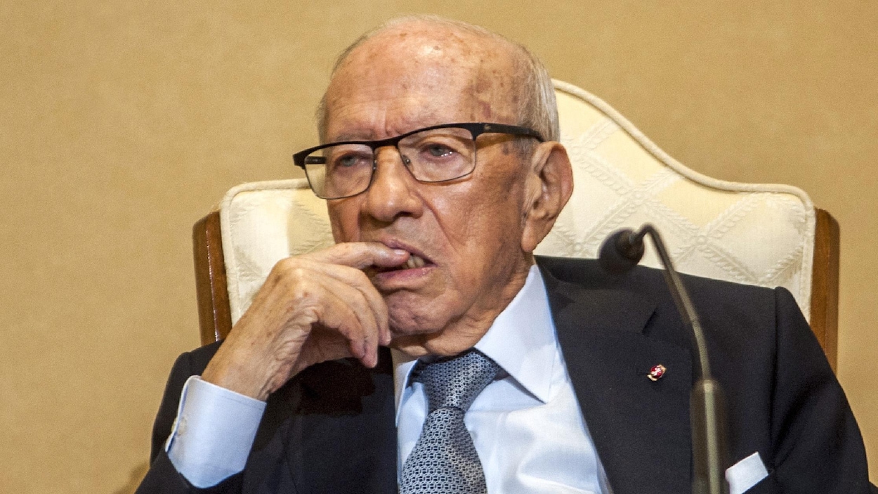 Il est assis dans un fauteuil et porte son index droit à sa bouche.