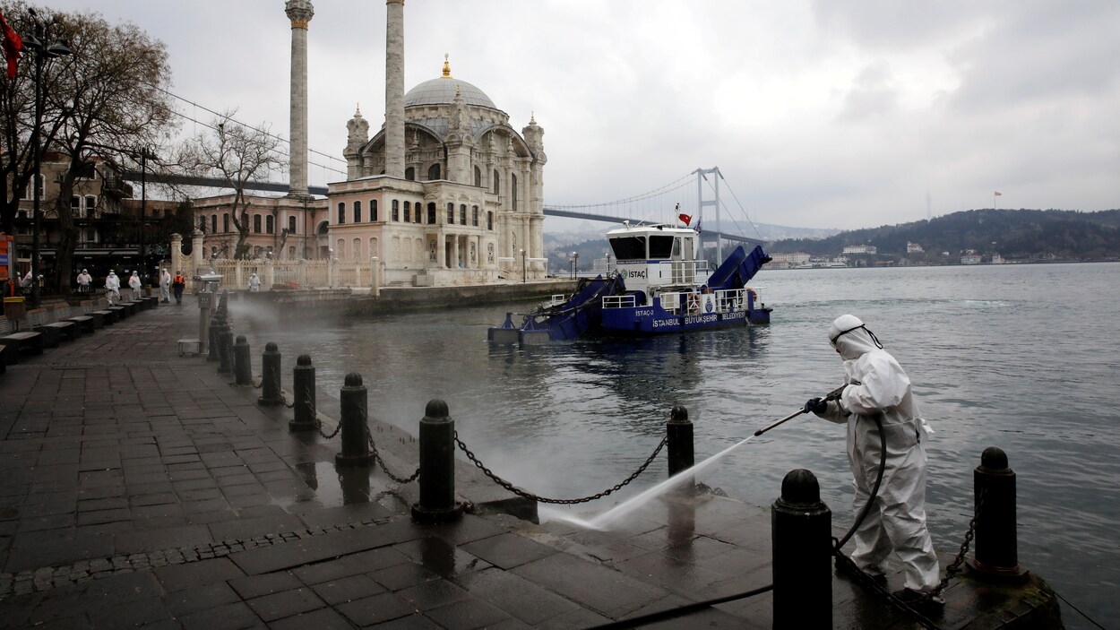 Un employé revêtu des pieds à la tête d'une camisole de protection vaporise du désinfectant sur le pavé, au bord de l'eau, avec une mosquée en fond de décor.