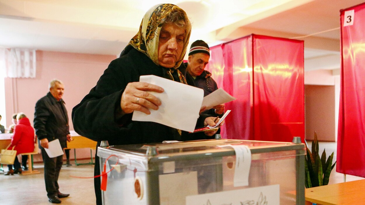 Une femme portant un foulard sur la tête et un manteau noir dépose un bulletin de vote dans une boîte dans un bureau de scrutin en Ukraine.