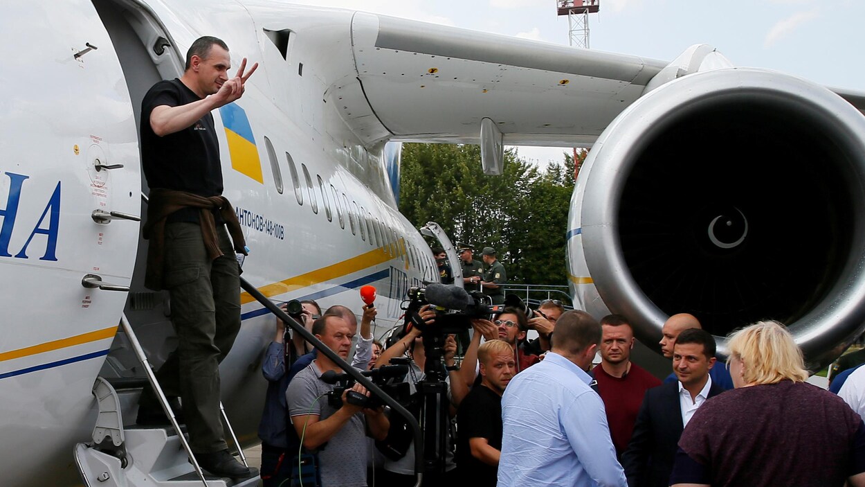 Oleg Sentsov fait un signe de la victoire avec sa main droite alors qu'il descend les escaliers de l'avion devant une foule et des caméras.