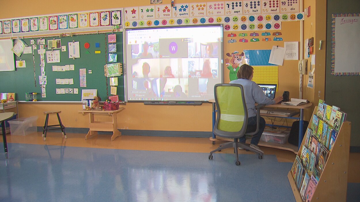 Une enseignante assise devant un ordinateur dans sa classe; au mur, un écran sur lequel est projetée une vidéoconférence, avec sa mosaïque de participants.