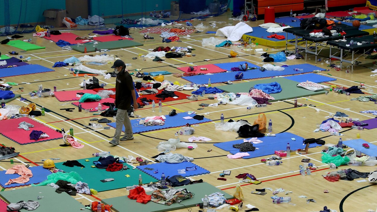 Une personne masquée marche dans un gymnase au plancher couvert de petits matelas, de vêtements et de babioles du quotidien.