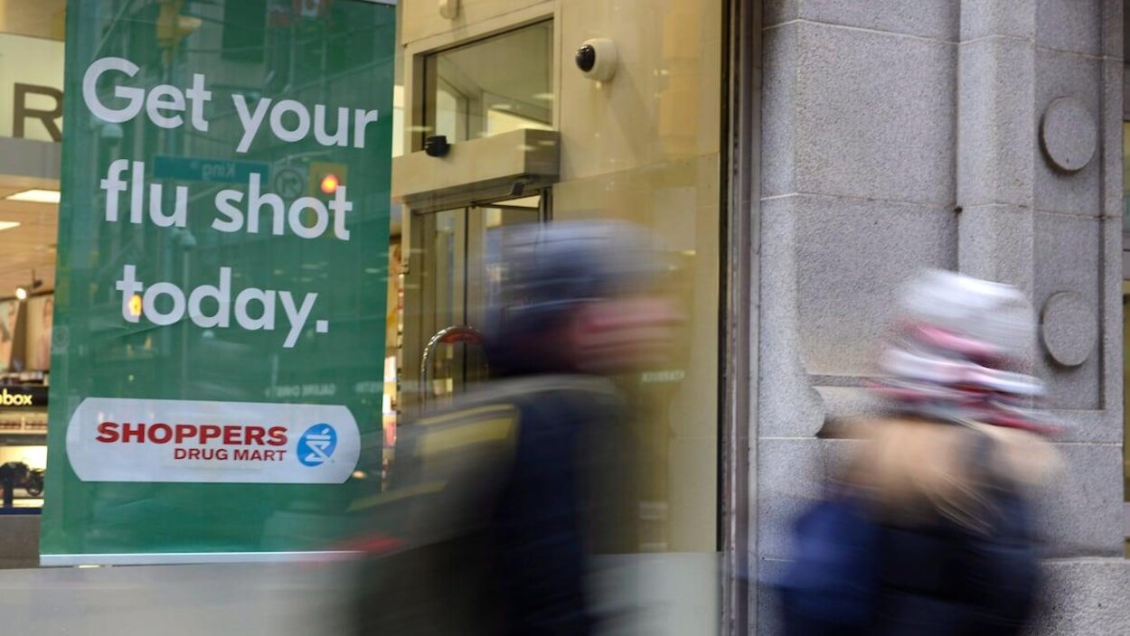 Une affiche dans une vitrine de pharmacie annonçant la disponibilité du vaccin contre la grippe, des gens marchent devant.