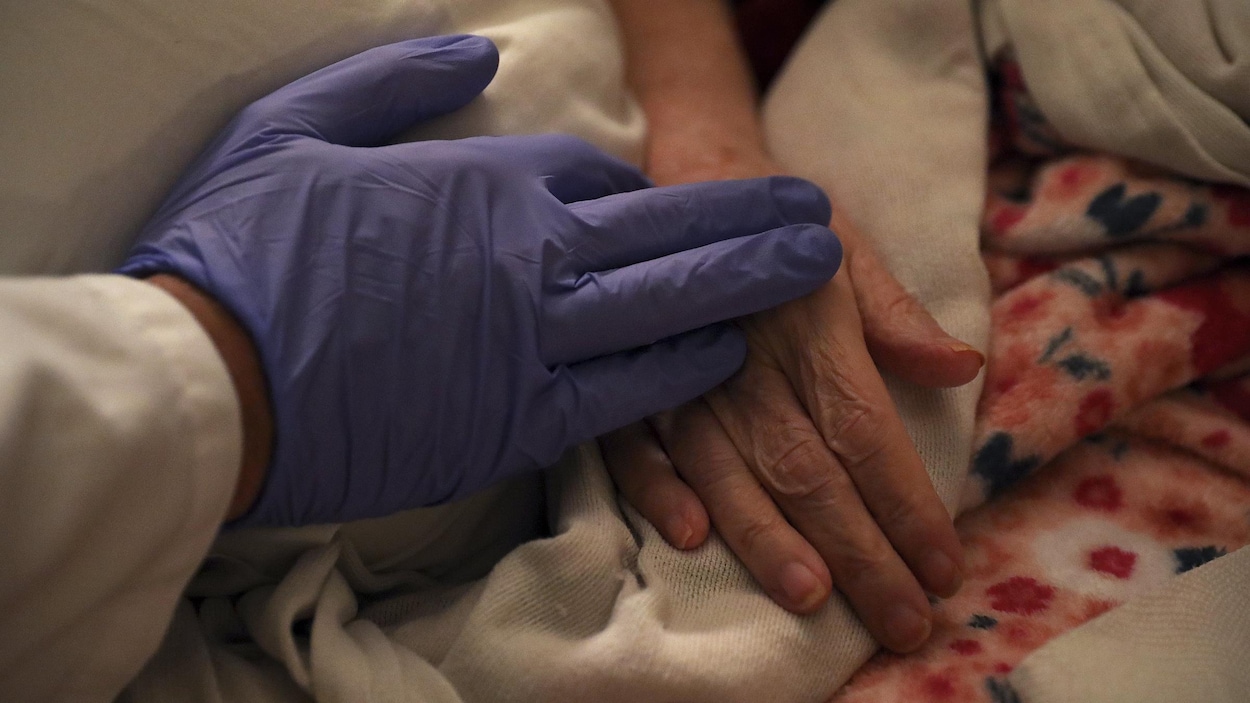 La main, couverte d'un gant médical bleu, d'un infirmier se pose sur la main d'une femme âgée couchée dans un lit.