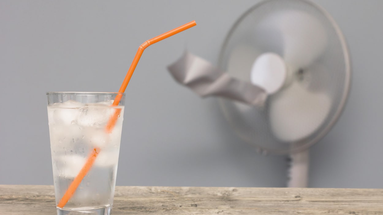 Verre d'eau glacée avec paille sur une table en bois et ventilateur de la maison en arrière-plan.