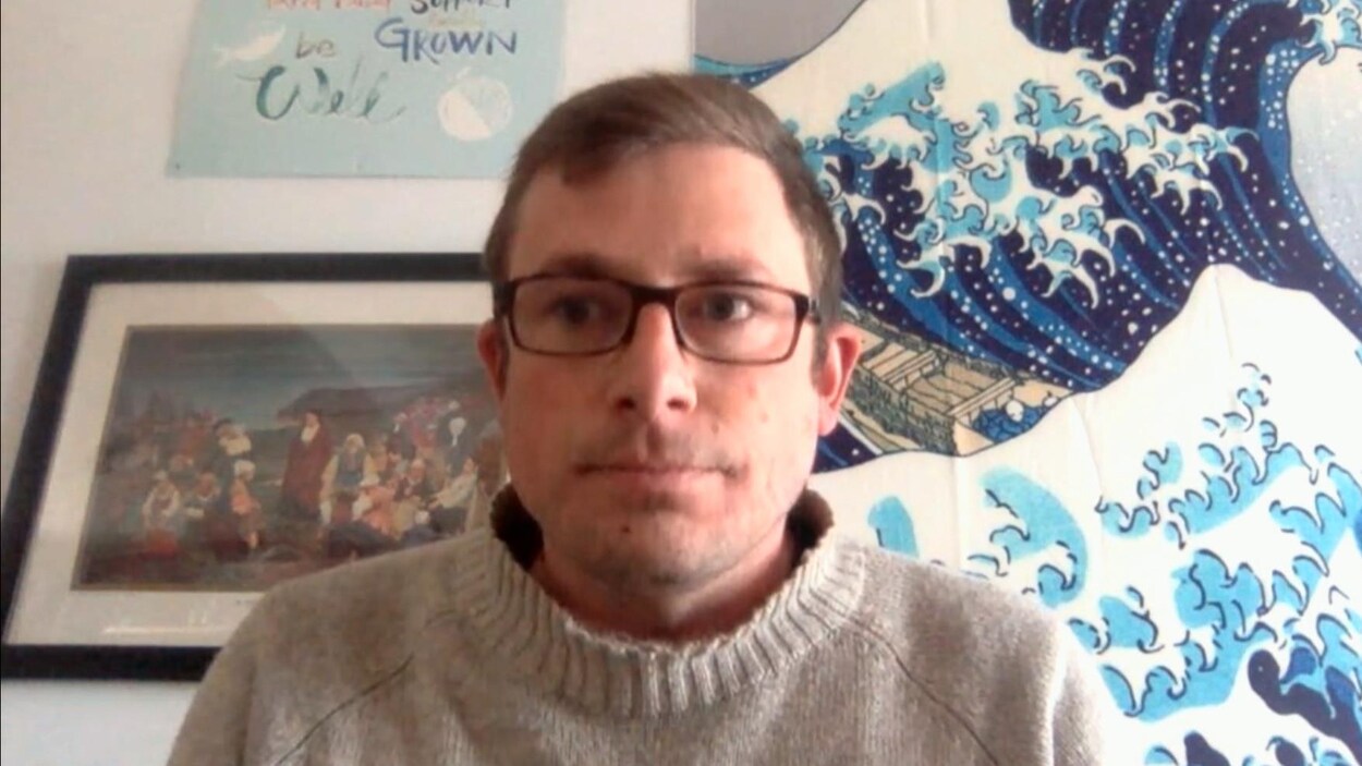 Une image d'un homme avec des lunette, avec une toile d'ocean derrière lui et un tableau de la déportation des acadiens.