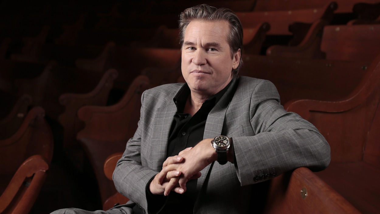 Val Kilmer est assis sur un banc en bois.