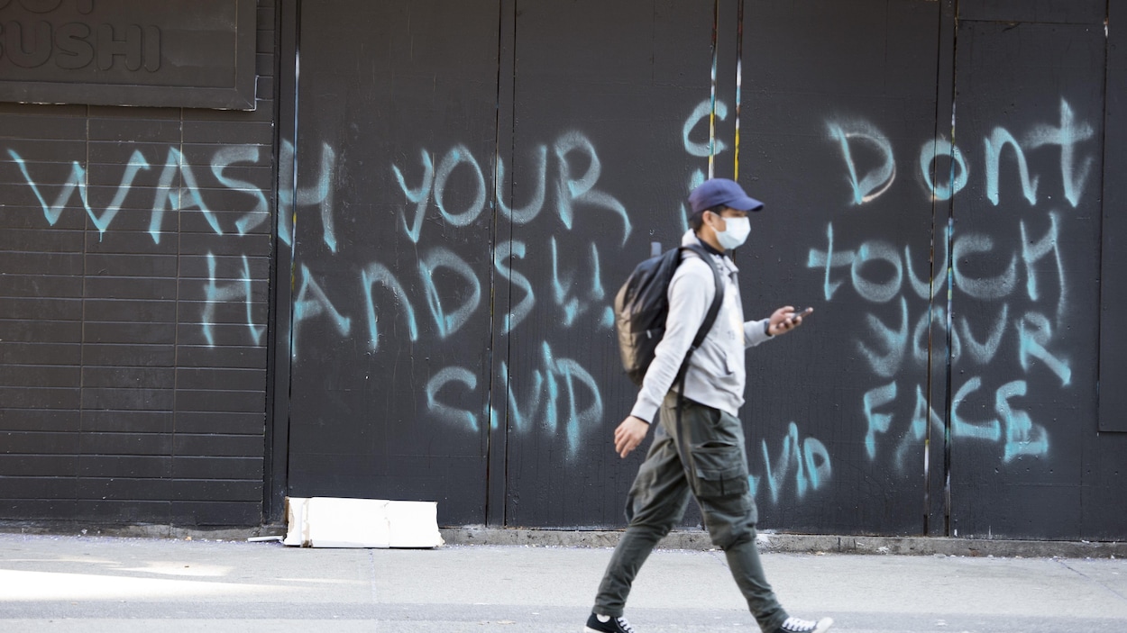 Un homme portant un masque passe devant un mur où on peut lire : « Lavez vos mains et ne touchez pas votre visage ».