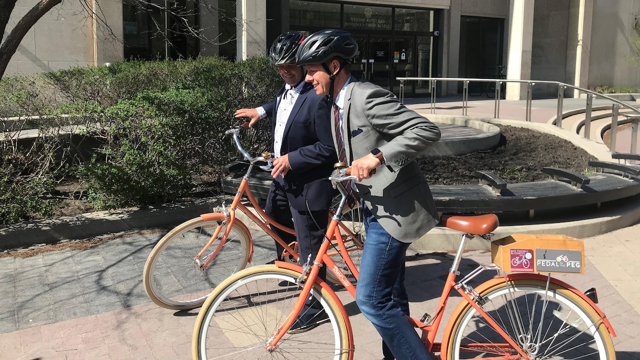 Deux hommes en costume s'élancent sur des vélos oranges devant un bâtiment municipal. Deux hommes en costume s'élancent sur des vélos oranges devant un bâtiment municipal.