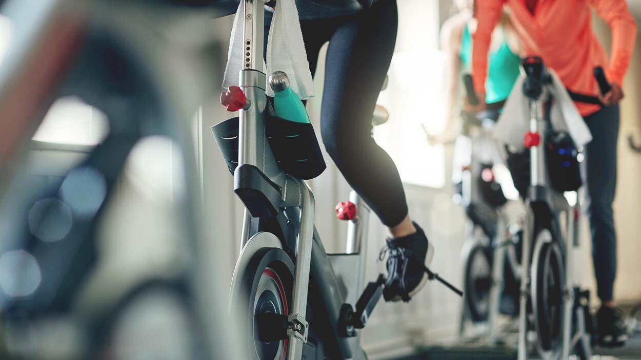 On voit des gens qui s'entraînent sur des vélos stationnaires. L'image est focalisée sur leurs jambes en action (on ne voit pas les visages).
