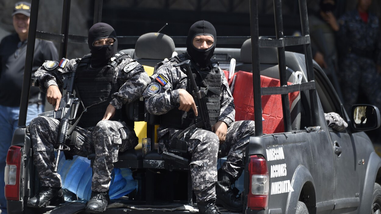 Deux hommes en uniforme, le visage masqué et portant des fusils automatiques, sont assis dans la boîte arrière d'une camionnette.