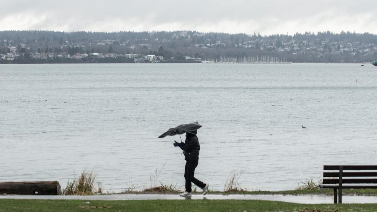 Une personne marche sur le bord de l'eau avec un parapluie soufflé par le vent.