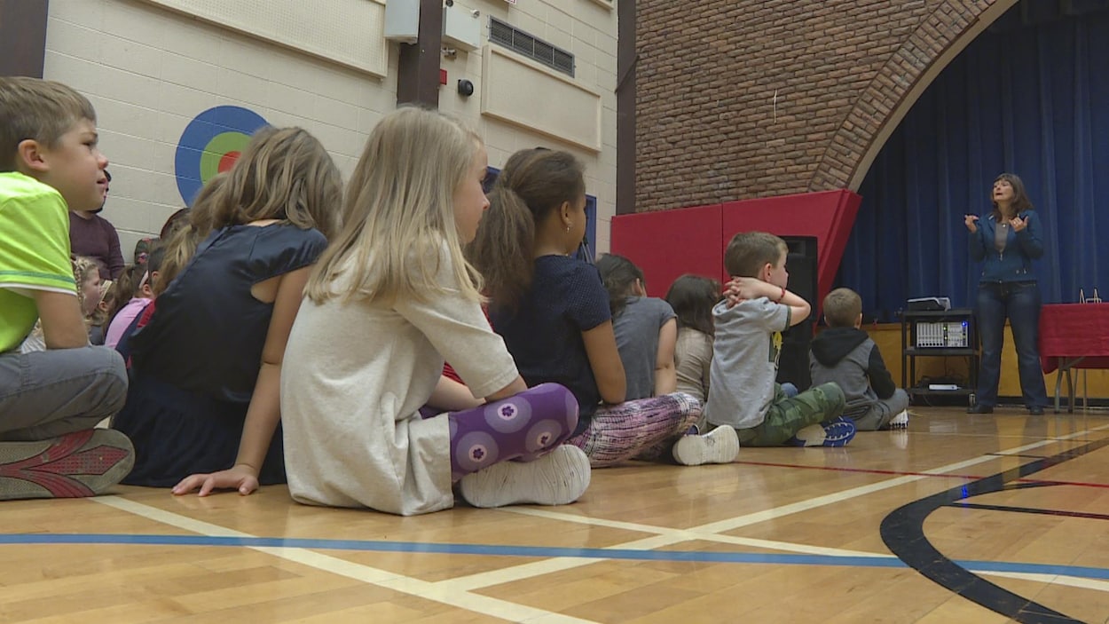 Des enfants sont assis dans un gymnase et regardent une femme qui leur parle à l'avant de la salle.
