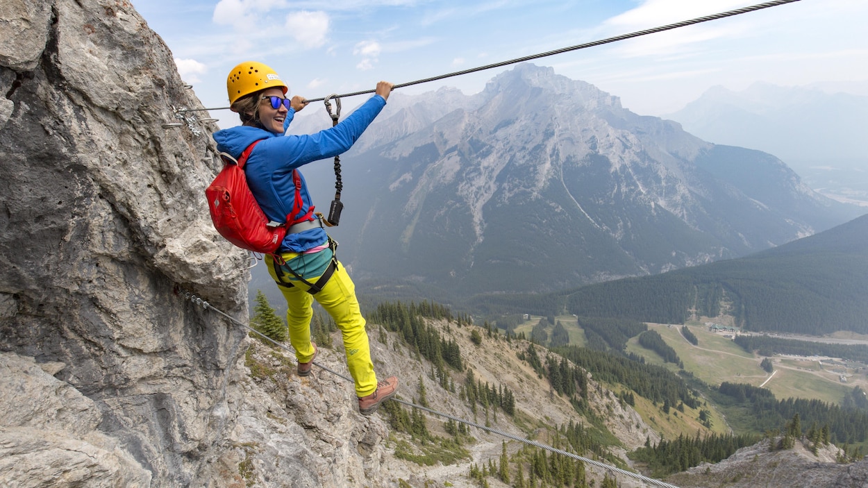 Une femme emprunte une passerelle en haute altitude. En arrière-plan, des montagnes rocheuses.