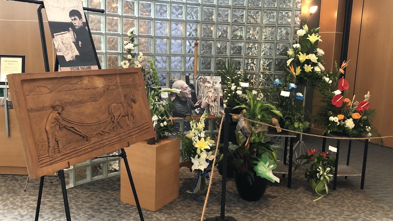 Plusieurs photos et oeuvres de Victor Dallaire entourées de fleurs dans un salon funéraire.