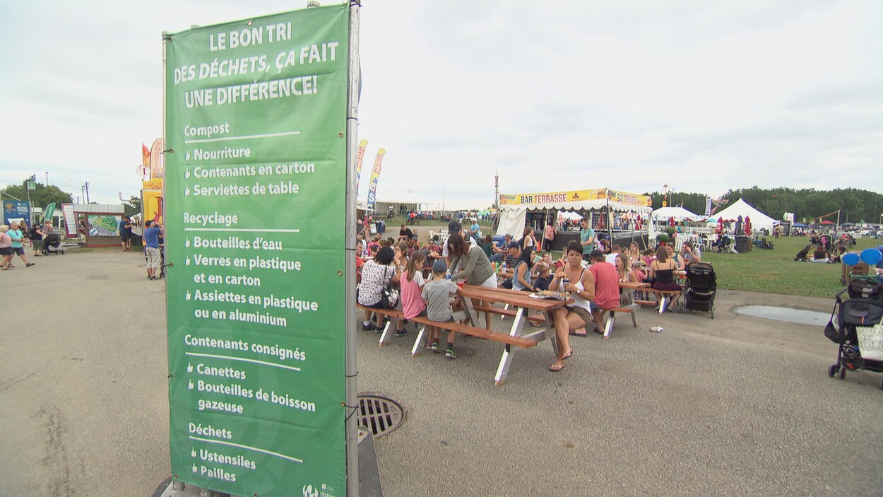Un panneau informatif sur les choses à jeter ou recycler devant plusieurs tables à pique-nique où mangent des festivaliers.