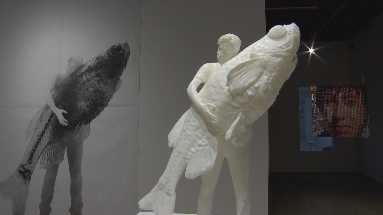 Une sculpture d'un jeune homme, qui tient dans ses mains un poisson de sa taille.
Derrière, dans l'ombre, une installation composée de petites bouteilles de verre colorées, qui forment le visage d'une jeune fille. Une sculpture d'un jeune homme, qui tient dans ses mains un poisson de sa taille.
Derrière, dans l'ombre, une installation composée de petites bouteilles de verre colorées, qui forment le visage d'une jeune fille.