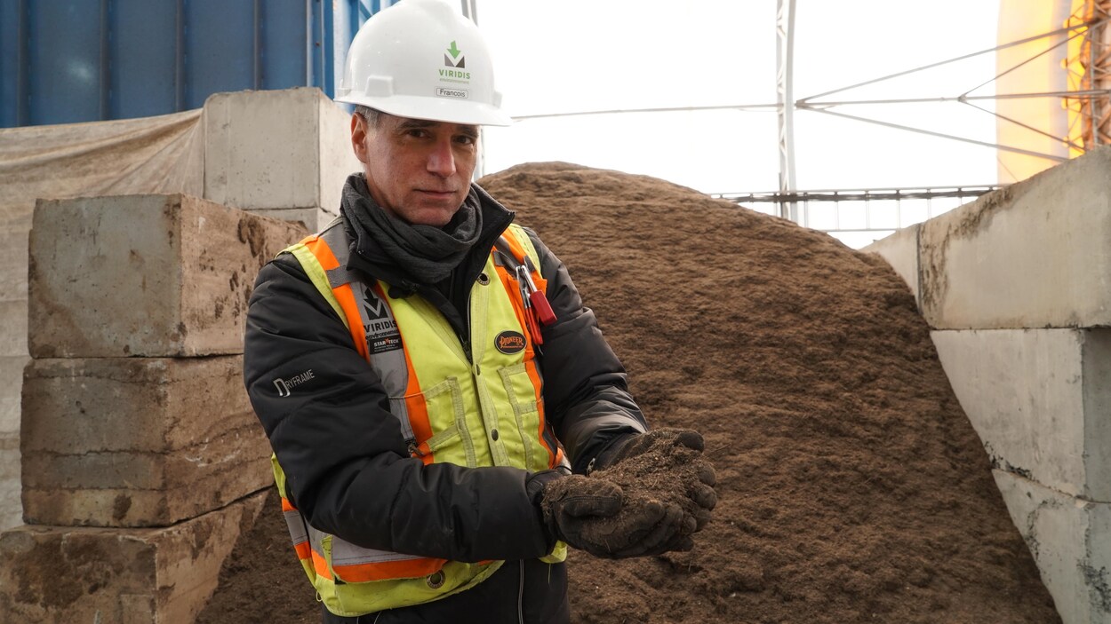 Un homme avec une veste jaune fluorescente et un casque blanc tient du compost dans ses mains.
