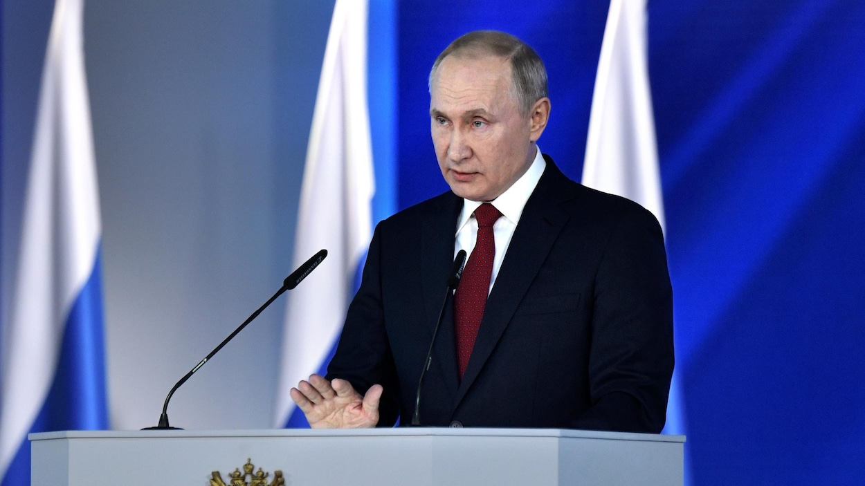 Vladimir Poutine parle derrière un lutrin, plusieurs drapeaux russes se trouvent derrière lui.