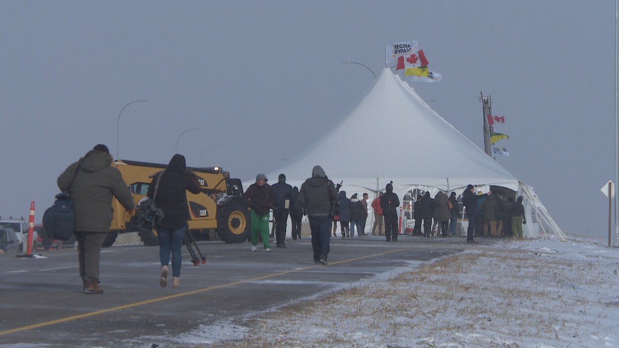 Des personnes se dirigent vers une tente installée sur une autoroute. Un engin de chantier est garé à proximité. Le temps est hivernal.