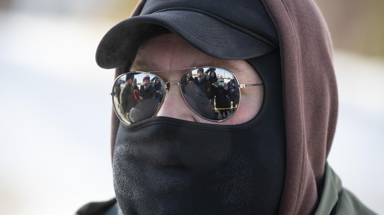 Plan rapproché d'un homme cagoulé dont les lunettes reflètent les journalistes devant lui.